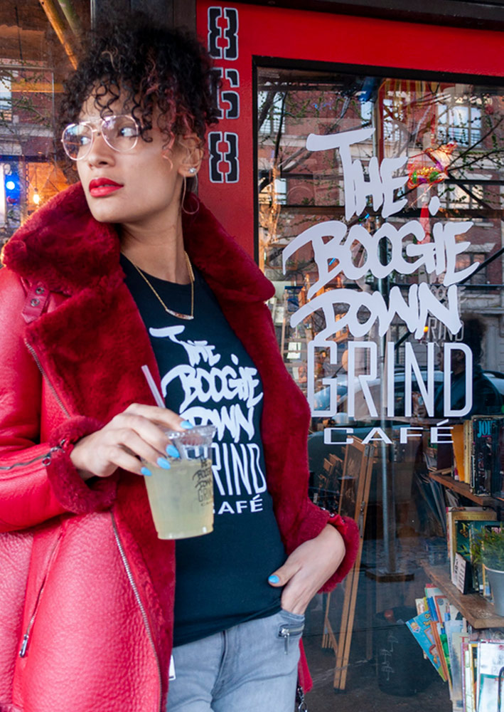 woman stands outside The Boogie Down Grind Cafe with drink