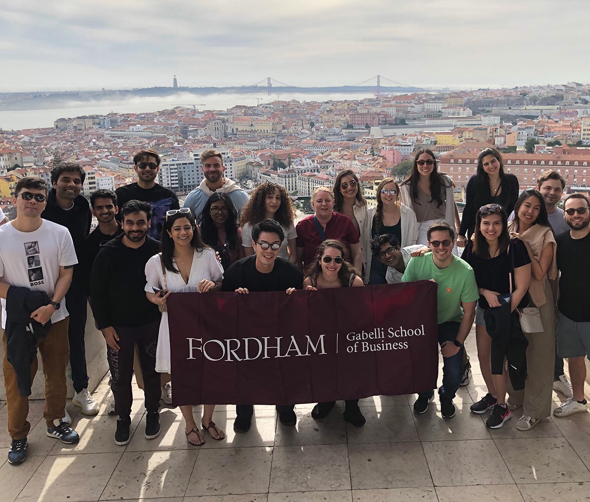 Full-time MBA students from Fordham Gabelli School of Business visited agricultural leaders, fintech firms, and business startups in Portugal as they all gather together outside smiling and posing for a picture while a few individuals in the front hold a dark burgundy Fordham Gabelli School of Business banner flag.