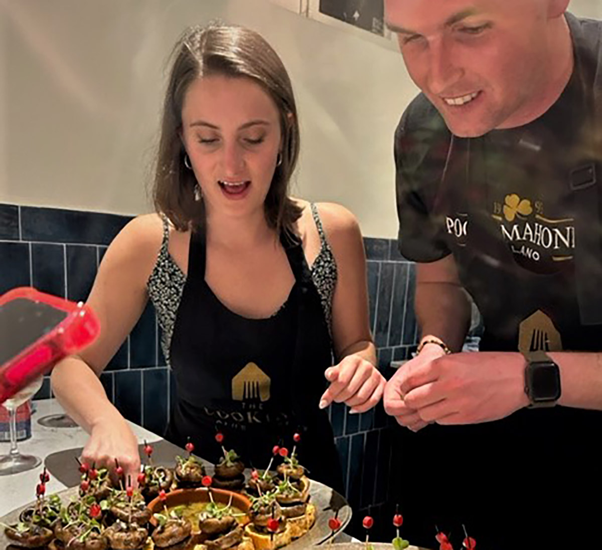 Along with a packed agenda of 14 company visits in Madrid, MBA students from Fordham Gabelli School of Business enjoyed a tapas cooking class as a woman and a man have aprons on learning how to make tapas next to each other as they glance downward at the food dishes.