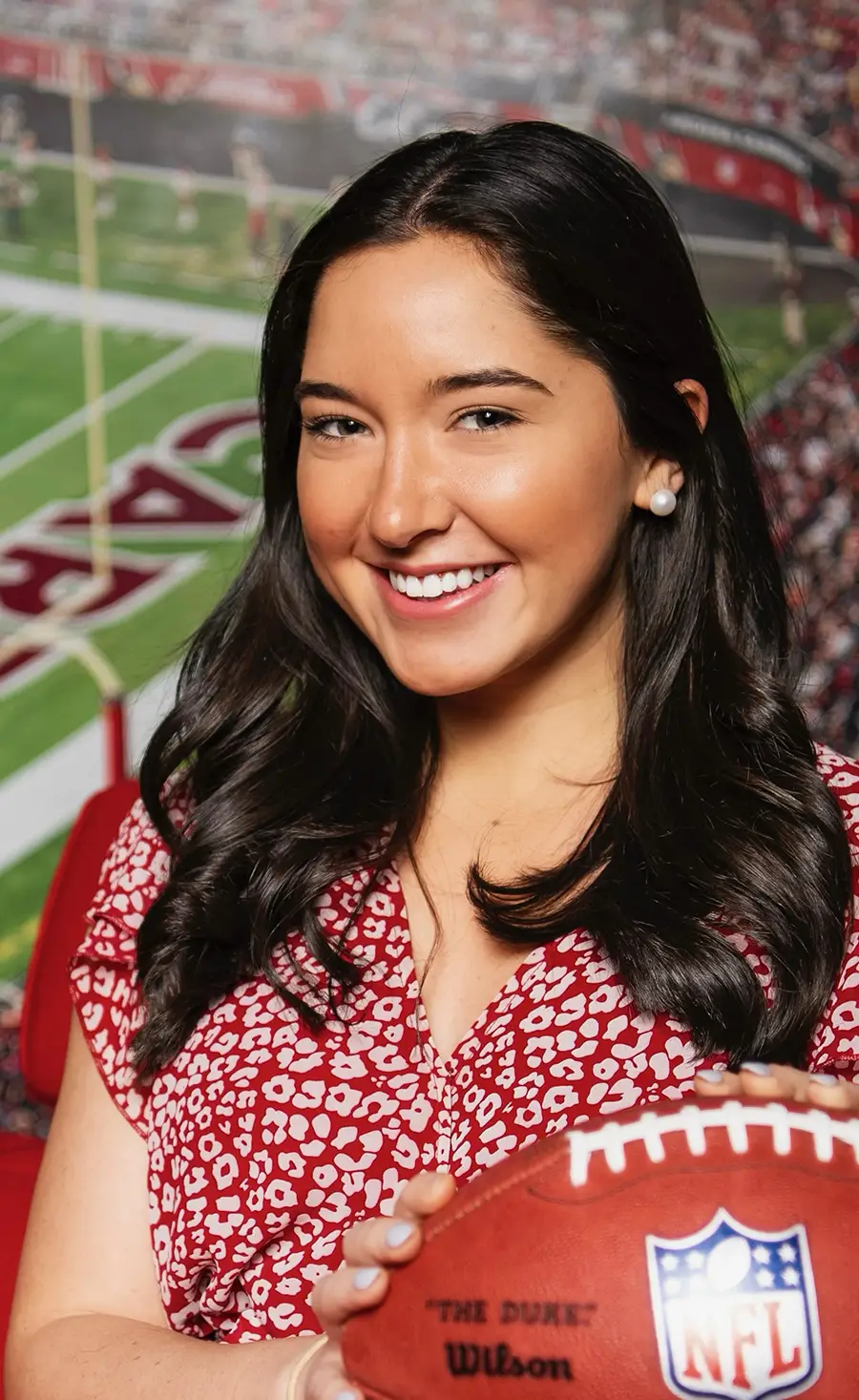 Portrait photograph headshot of Elizabeth "Libby" Vernon, BS' 22 smiling in a red blouse top that has white small custom flower shape patterns all over as she holds a National Football League shaped game-day ball (foreground) while in the background behind her is a zoomed in wall picture perspective of the Arizona Cardinals stadium field area