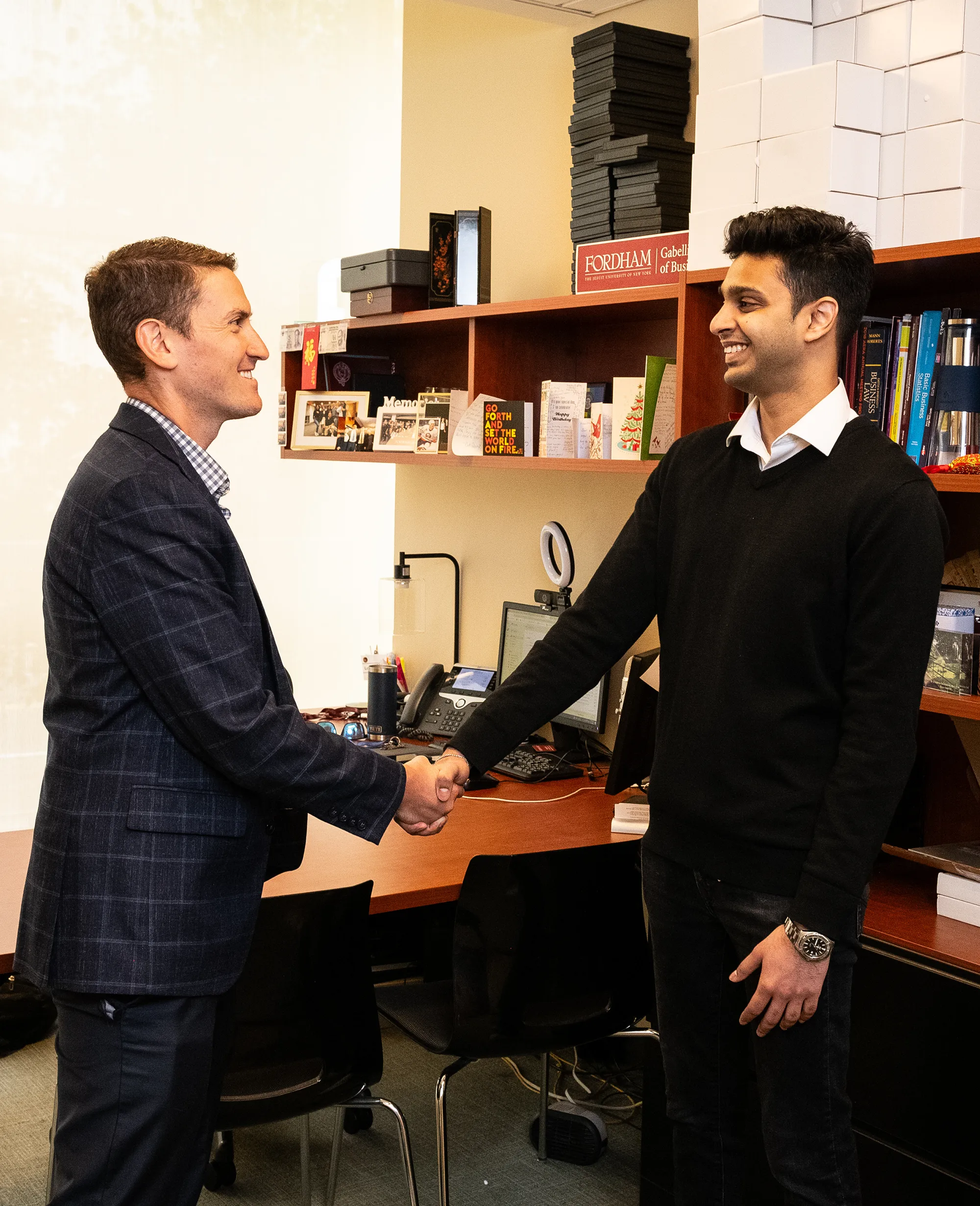 A professional setting where two men shake hands in an office, smiling at each other. One is wearing a dark blazer, while the other is in a black sweater. A "Fordham Gabelli School of Business" sign is visible on the office shelf behind them.