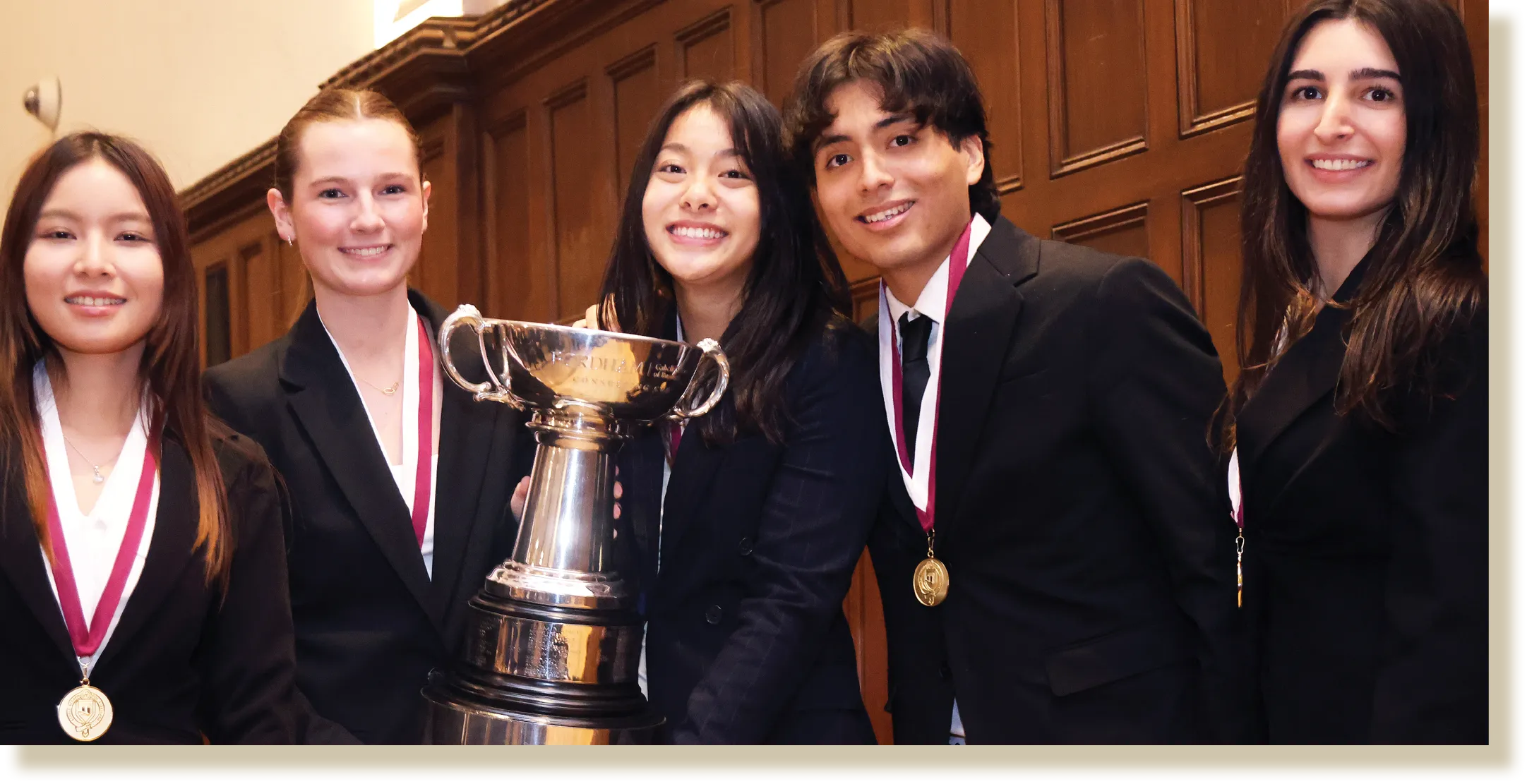 A group of five students dressed in formal business attire, smiling and posing with a large silver trophy in a wood-paneled room.