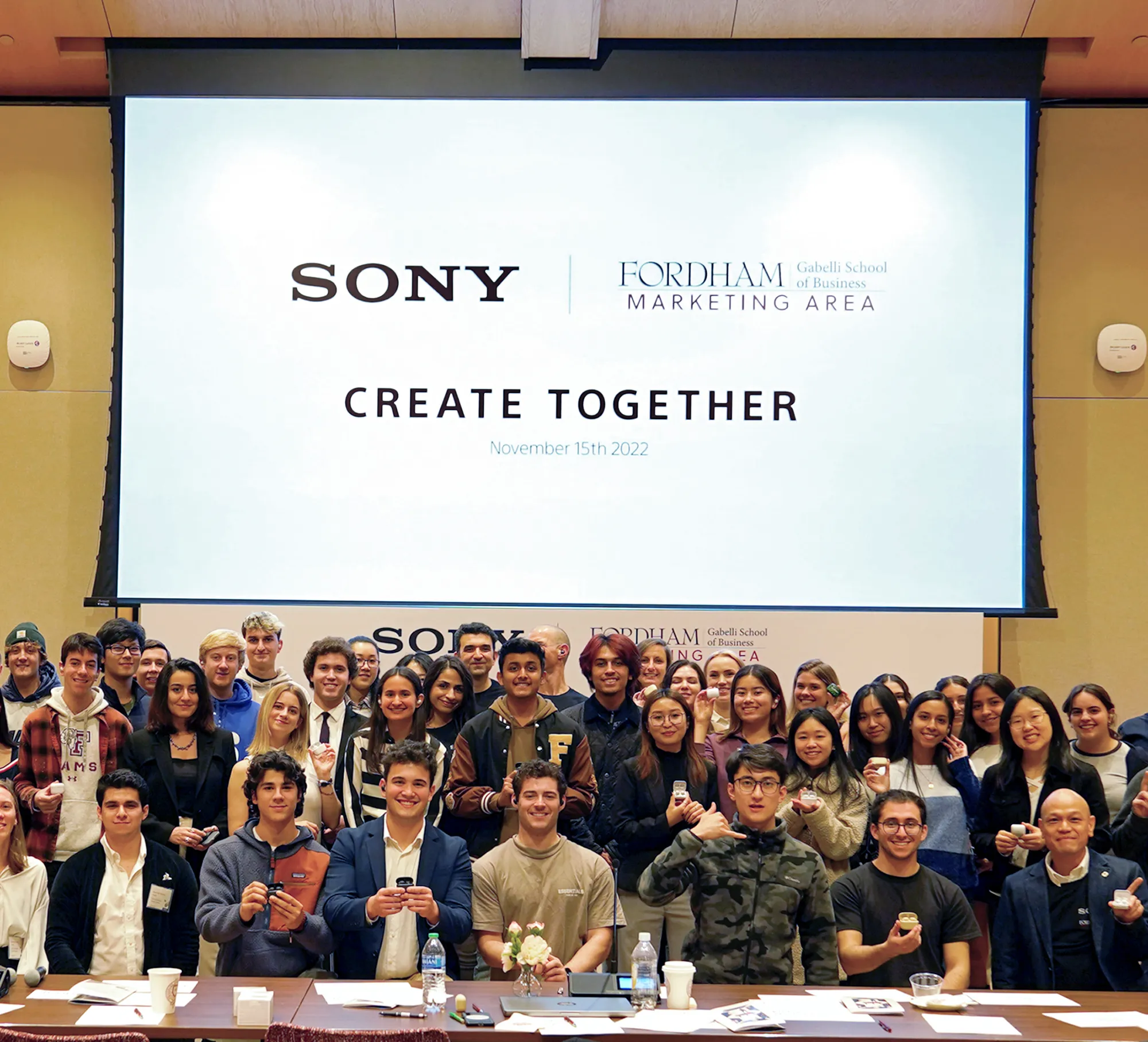 A large group of students and faculty members posing in front of a projector screen displaying "Sony | Fordham Gabelli School of Business Marketing Area - Create Together" during a conference event.