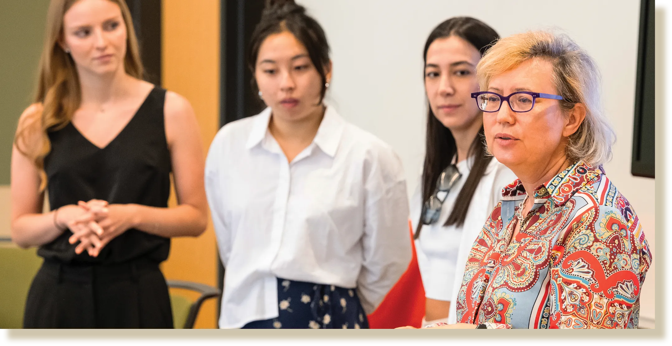A group of four women standing in a classroom setting, with one woman wearing glasses and a colorful blouse speaking while the others attentively listen.