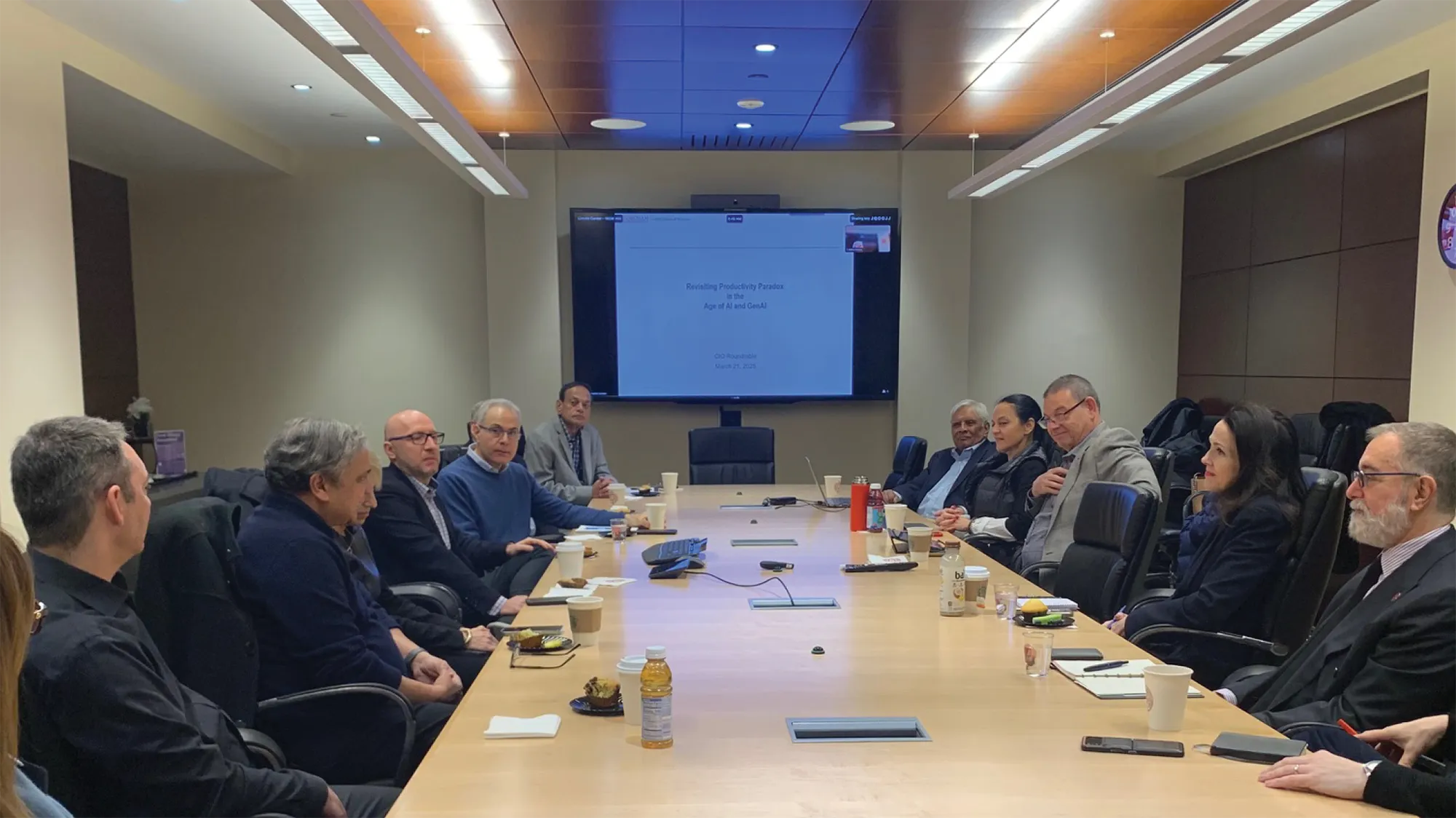 A group of people seated around a long conference table in a modern meeting room, participating in a roundtable discussion. A large screen at the end of the room displays a presentation titled “Revisiting Productivity Paradox in the Age of AI and GenAI – CIO Roundtable – March 21, 2024.”