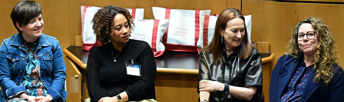 Four adult women are seated next to each other at the head of a large room, listening to a speaker.