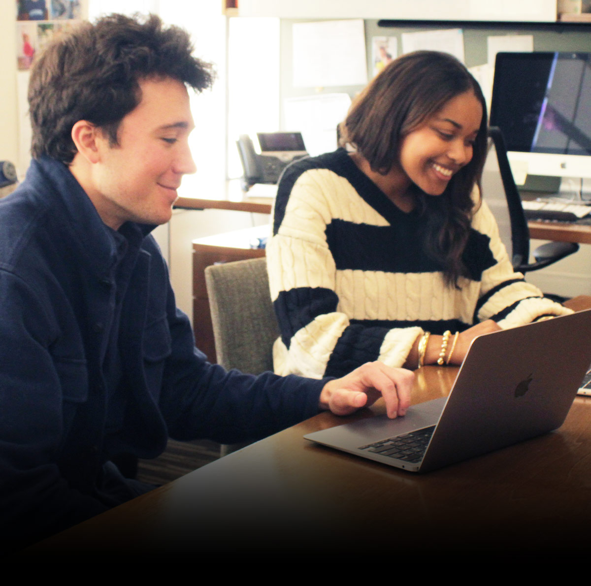 Two young adults are looking at a laptop on a desk and smiling. They are working together or enjoying something on the screen.