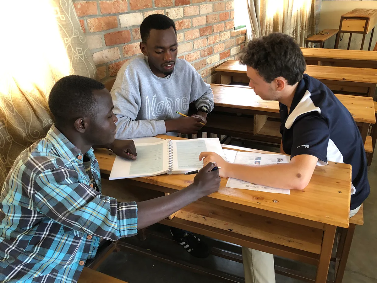 Three men, two in plaid shirts and one in a sweatshirt, sit around a wooden desk in a classroom setting with empty wooden desks behind them, looking down at open binders and papers.