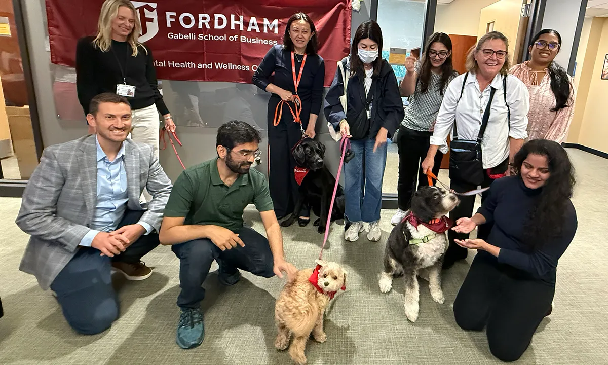 A group of nine people pose in a brightly lit hallway with three therapy dogs wearing red bandanas. A red banner is visible behind them.