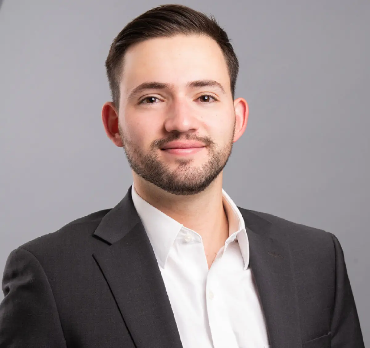 Headshot photograph of Mateo Ribadeneira, a man grinning in a black business blazer suit and white button-up dress shirt underneath