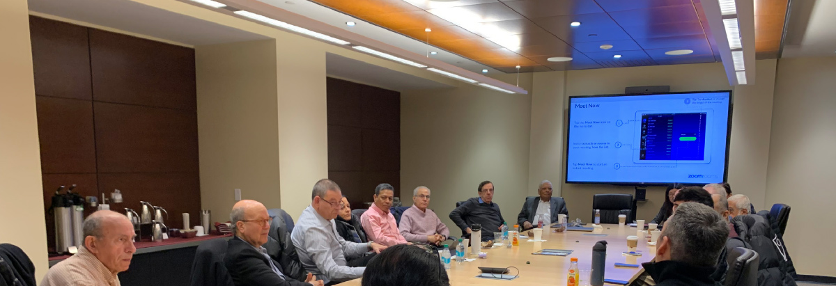 <br />
A large group of professionals sits around a long conference table during a roundtable discussion, with a Zoom Rooms presentation on a screen.